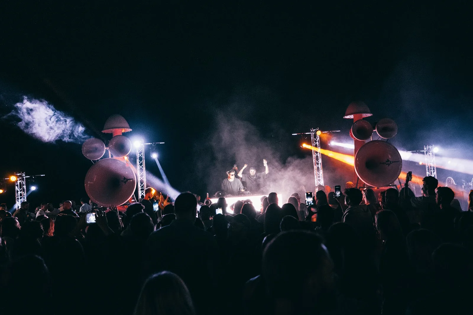 The stage at night seen from the crowd, two DJs under smoke and orange light
